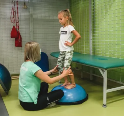 Fizjoterapeutka ćwiczy z dziewczynką stojącą na półkuli sensomotorycznej w sali ośrodka Troniny