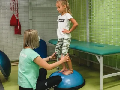 Fizjoterapeutka ćwiczy z dziewczynką stojącą na półkuli sensomotorycznej w sali ośrodka Troniny